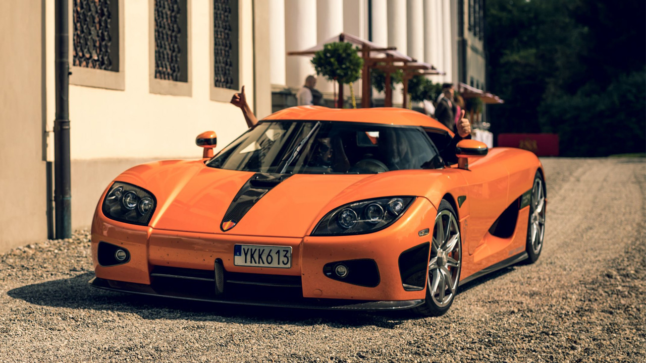passengers are thumbing up while sitting in orange colored Koenigsegg CCXR