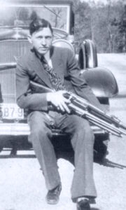 Clyde barrow sitting on a jeep and holding a gun