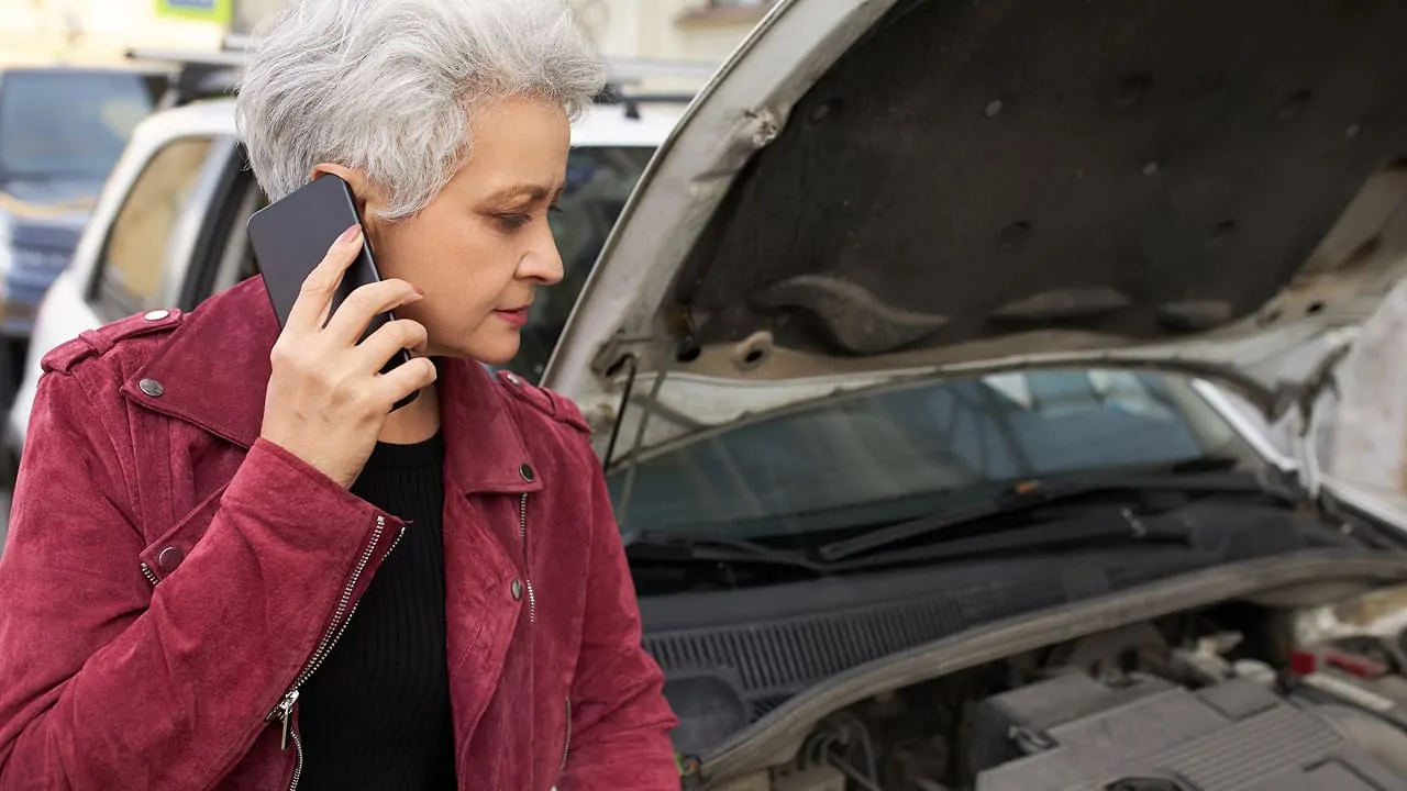 A grey-haired woman is standing next to her broken-down car