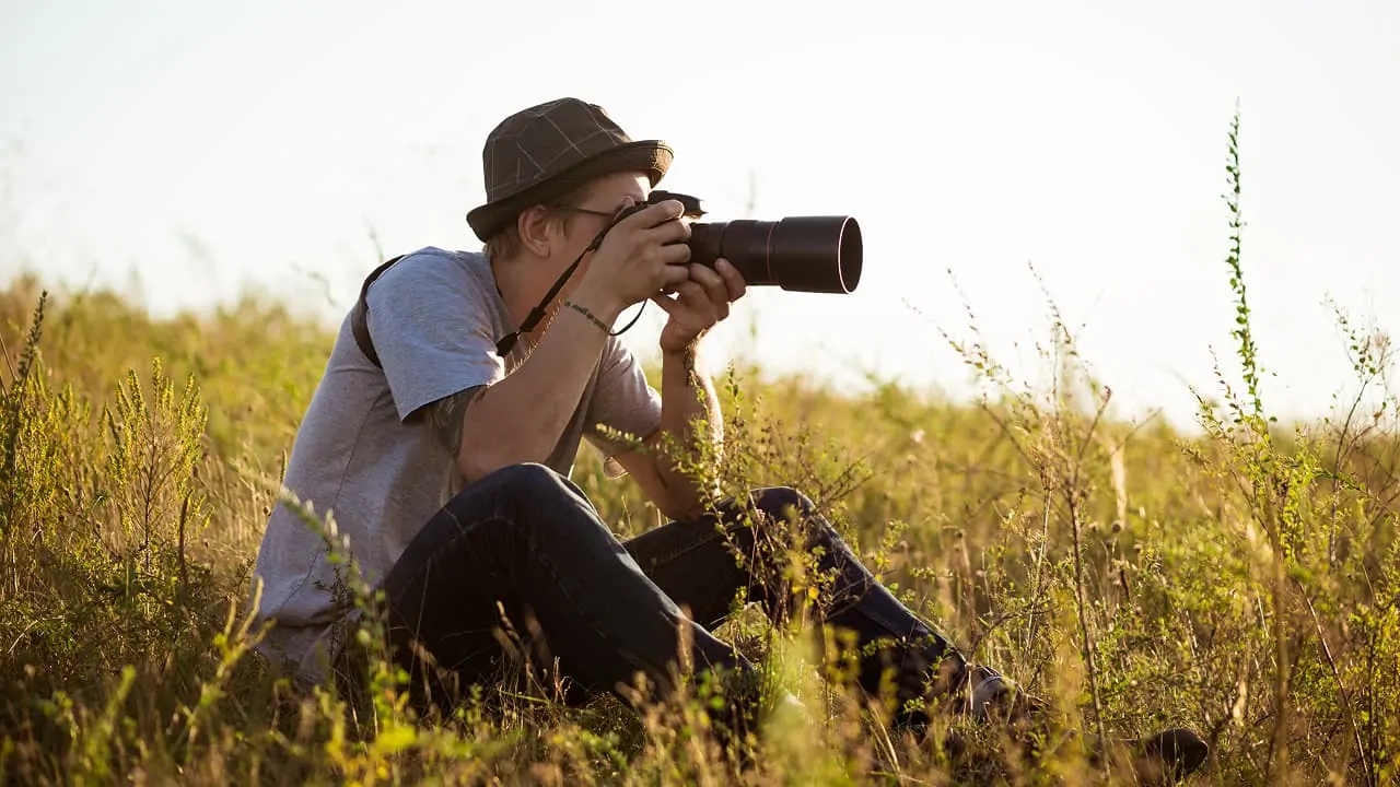A young male photographer is sitting in a field, taking pictures of wildlife
